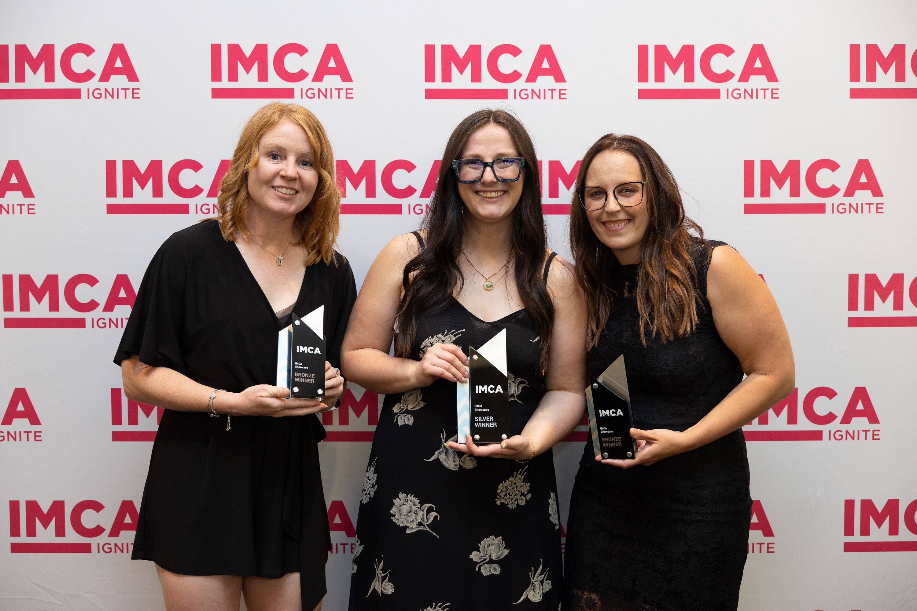 Three women smiling with IMCA backdrop, holding awards.
