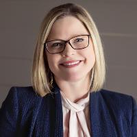 Professional woman on gray background with blonde hair and black glasses, wearing a black jacket