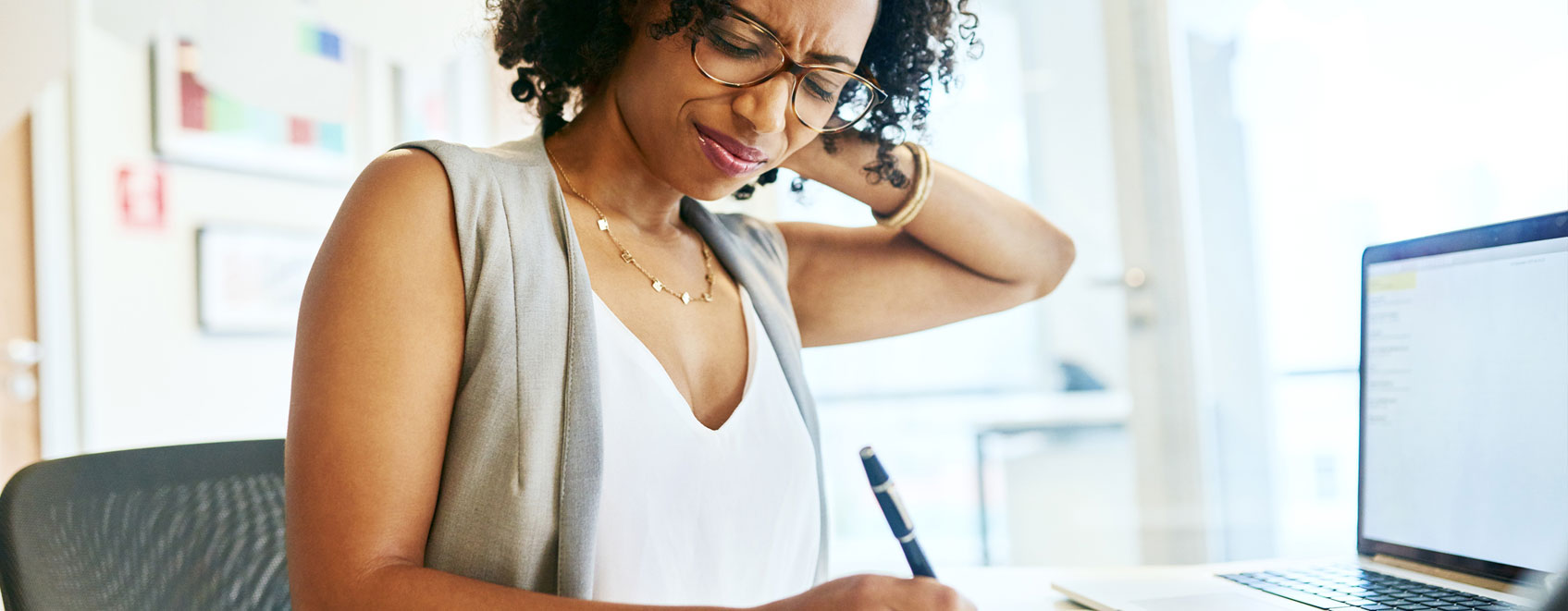 person at desk with laptop, wincing and rubbing back of their neck